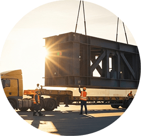 Crane lifting oversized steel structure onto a flatbed truck at sunset
